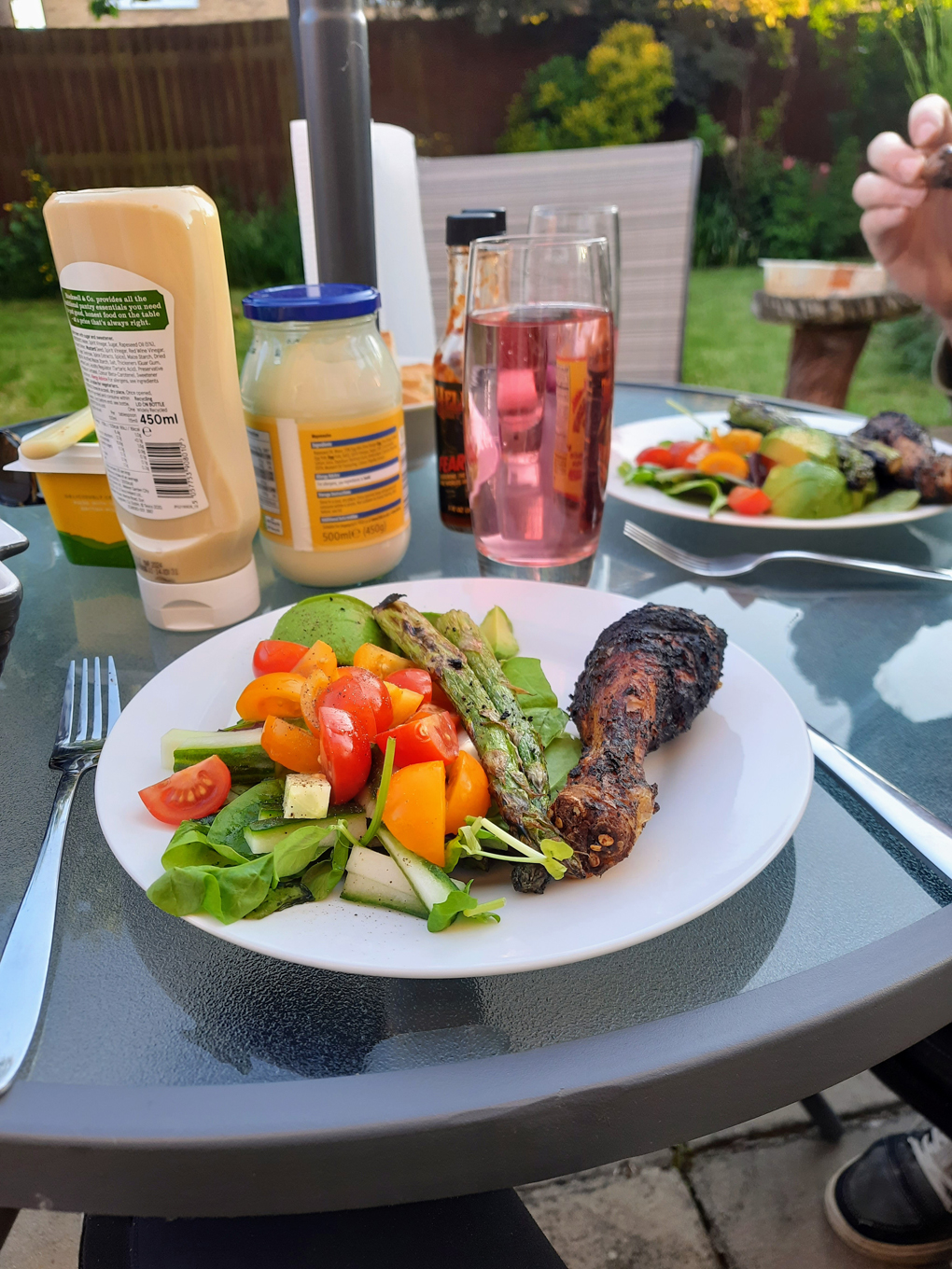Picture taken outdoors on a sunny day, a plate of fresh salad, and roasted veg and chicken is surrounded by a cool glass of water and condiments.