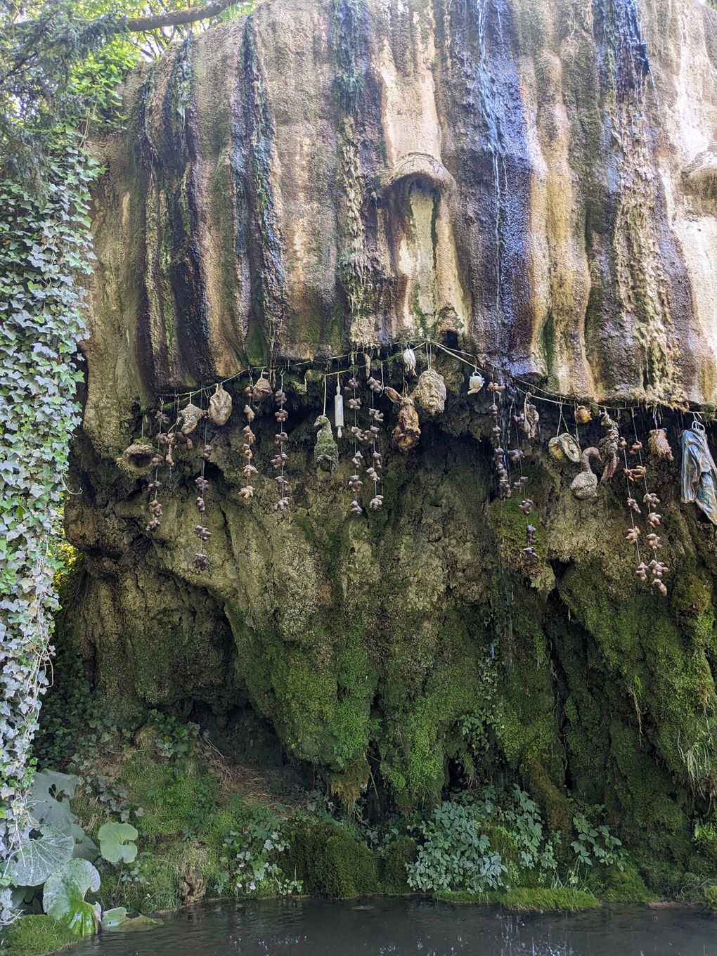 A rocky outcrop over which water drips into a pool below. A multitude of objects are hung in the path of the water as it drips down slowly turning them into stone including teddy bears, shoes, handbags and what looks like a human head