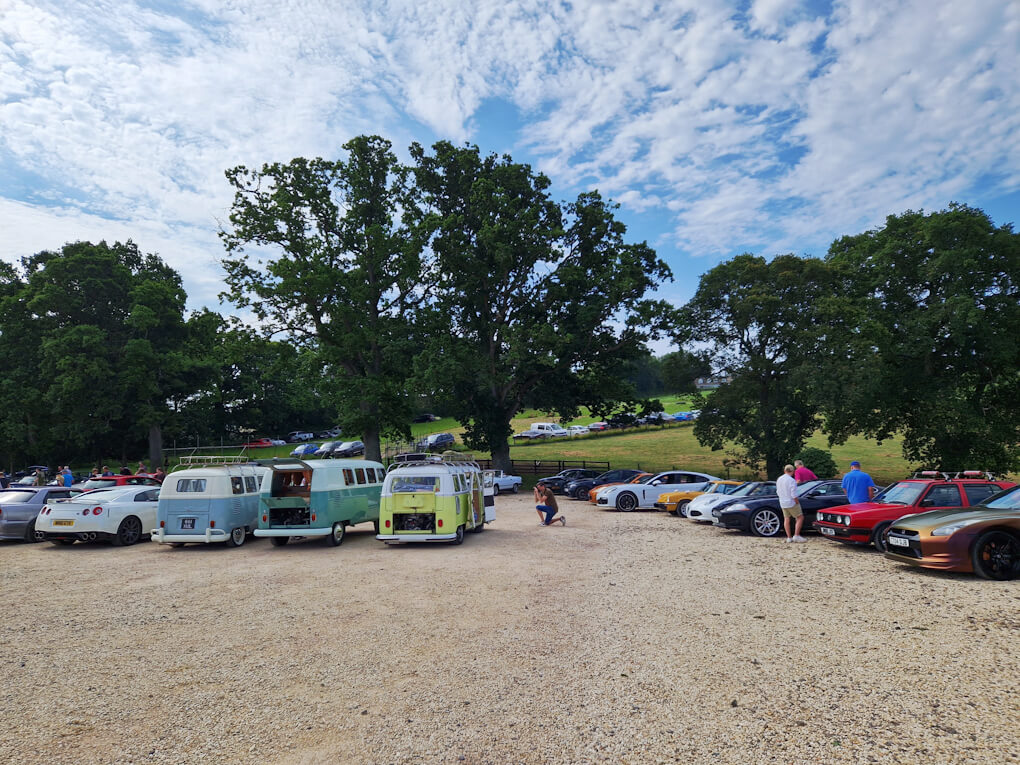 An eclectic collection of different cars and vans, parked in front of a green trees and hills