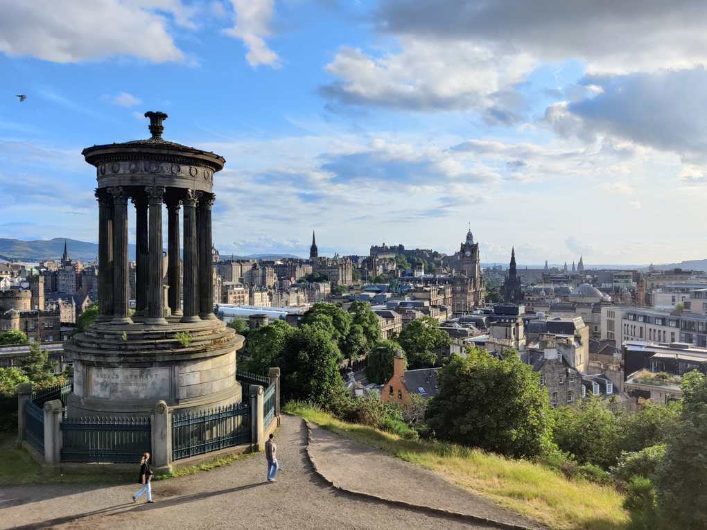 The view across the city of Edinburgh from Calton Hill on a clear summer day. On the left is a round building with columns, like a classical temple, in the distance is Edinburgh castle.