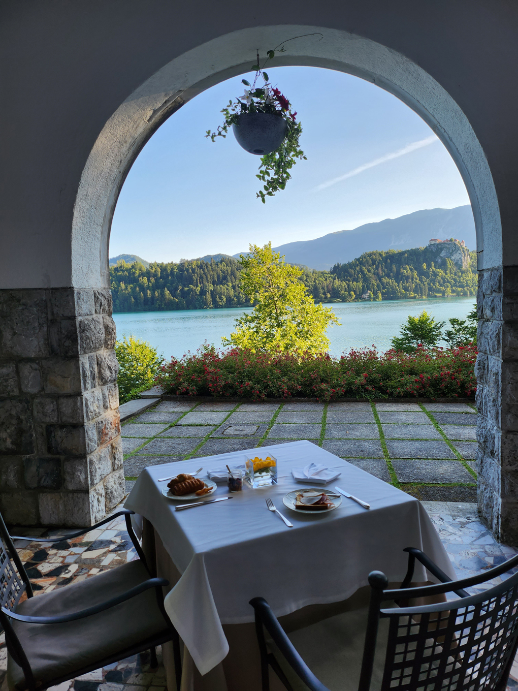 A table laid for breakfast is at the forefront just in front of an arch. Through the brick arch is a lake with an island. In the distance are mountains.