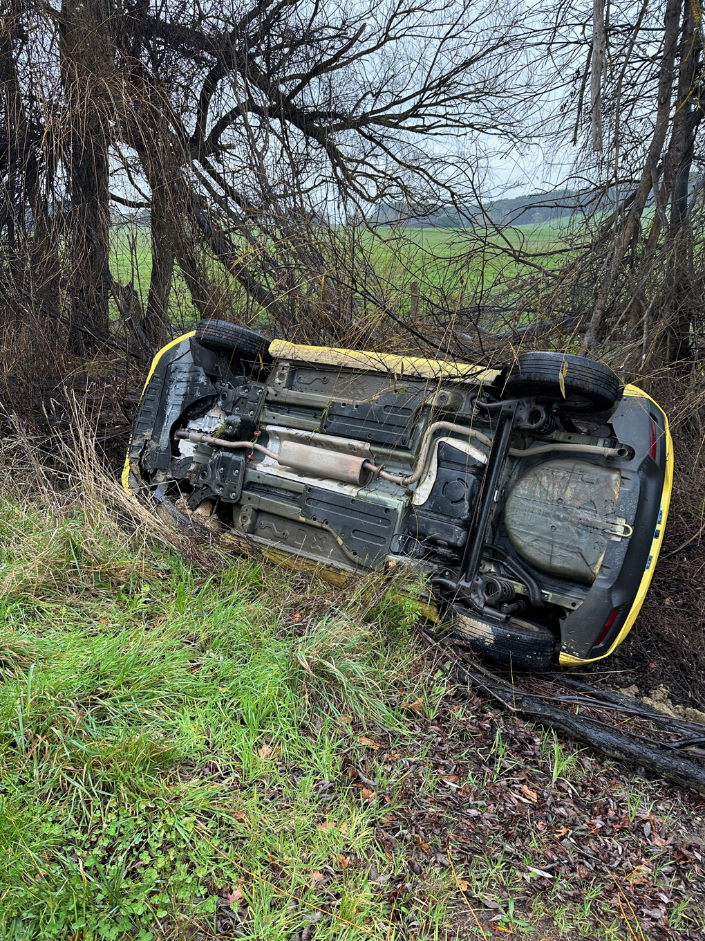 A rolled car off the road, on its right side with trees behind