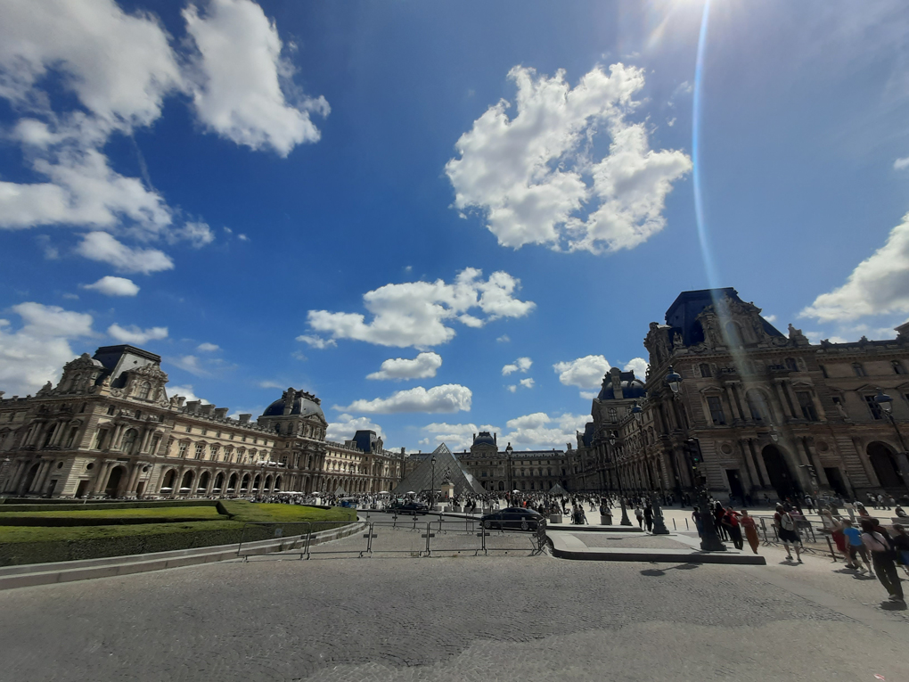 A wide view of the Louvre in Paris in the sunshine. The sky is vibrant blue with few clouds, the plaza is bustling with people, and the famous pyramid is in the centre.