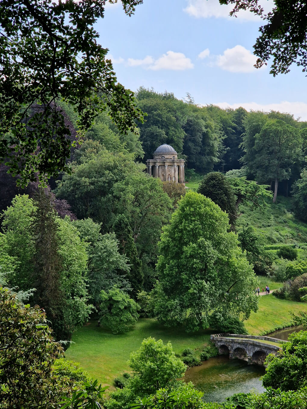 A lush green hillside covered in trees, with a sandstone temple near the top, and a multi-arch bridge over a shallow river at the bottom