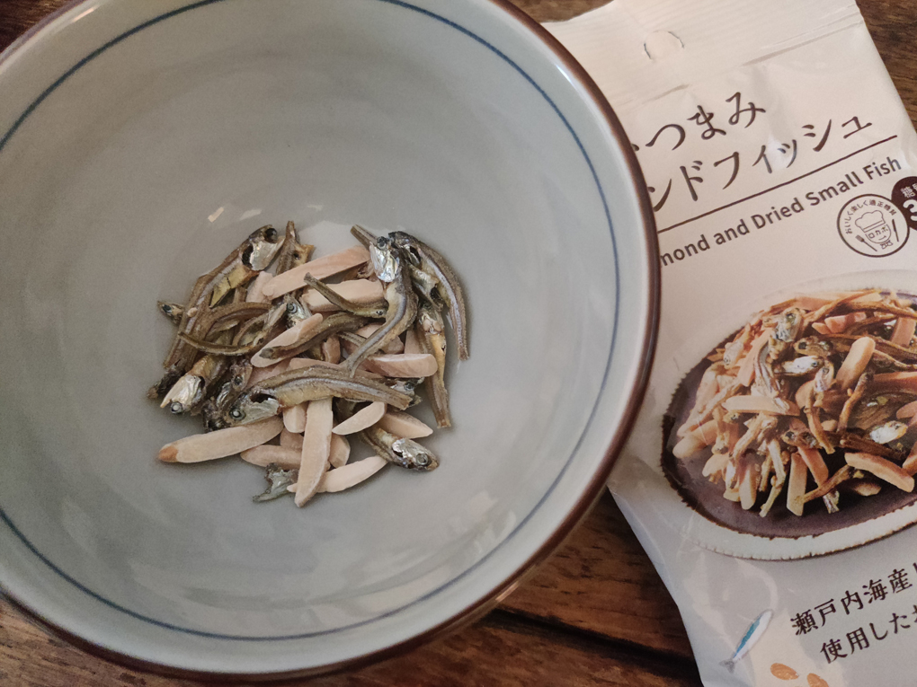 A small ceramic bowl contains a mix of tiny silver fish and flakes of toasted almond alongside is the shiny packaging covered with Japanese writing and a picture of the fish and almonds