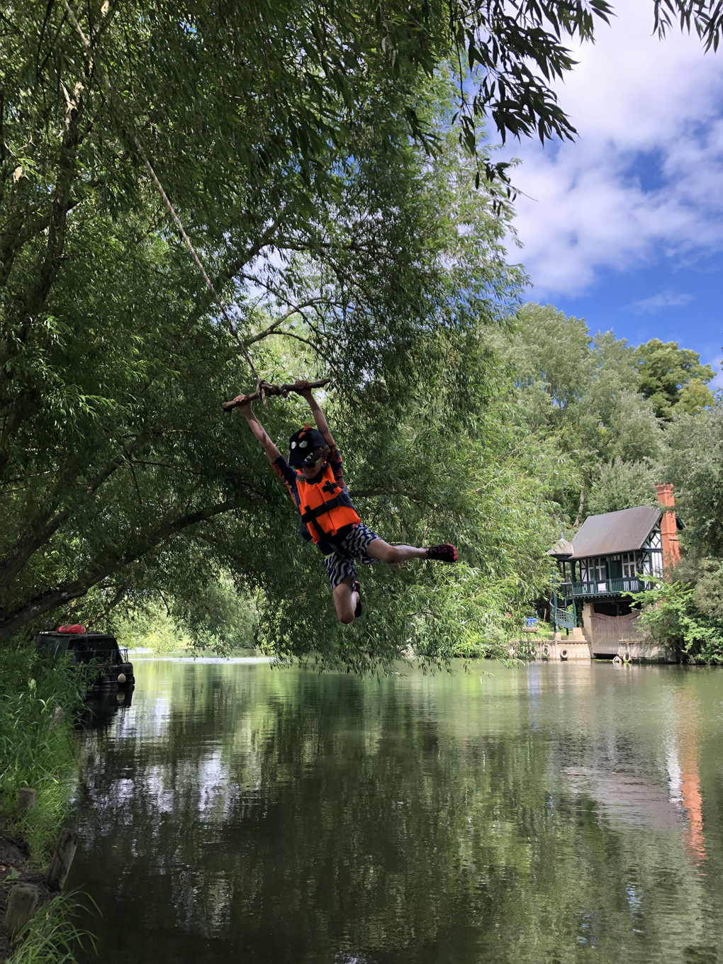 A small boy wearing shorts and a life jacket holding on to a rope swing, swinging out over the river thames