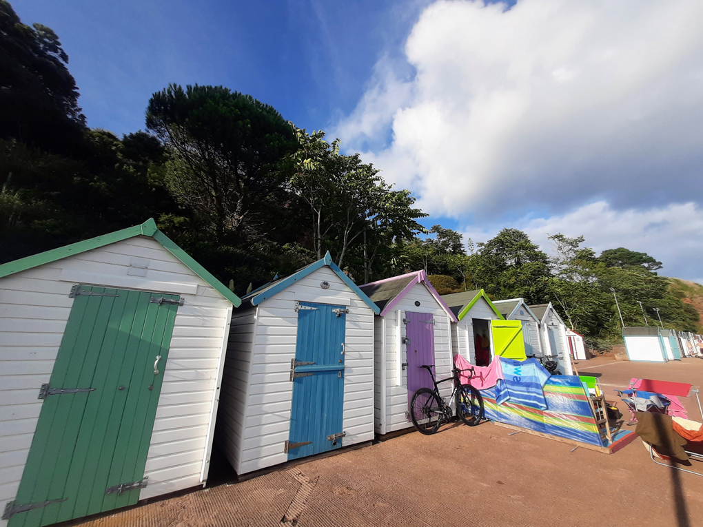 A row of brightly painted beach huts, white with blue, green, and purple doors all in a line. Outside one beach hut is a pedal bike and a windbreak flapping in the breeze.