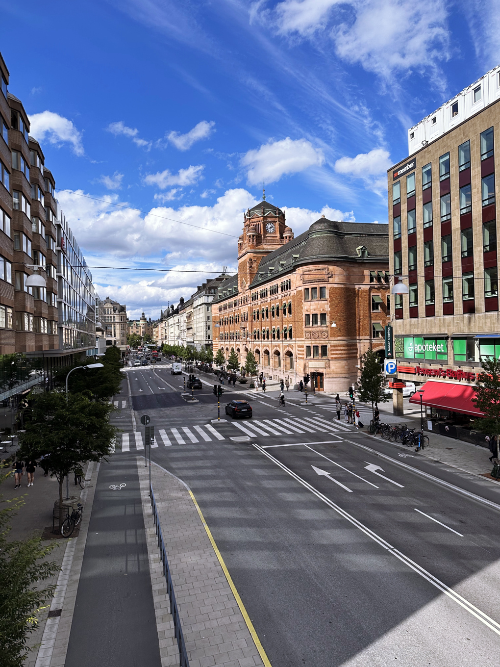 A straight, wide city road with little traffic, flanked by red, grey, and glass buildings of different architectural styles, under cloudy blue skies