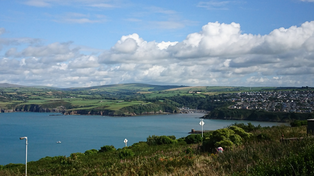 A view of Fishguard harbour with its lovely little town and small horse-shoe shaped marina on one of the few sunny days in August, under a rare blue sky with a bank of cumulus clouds along the horizon. A great view point at the top of a cliff on the opposite side of the harbour in Goodwick.