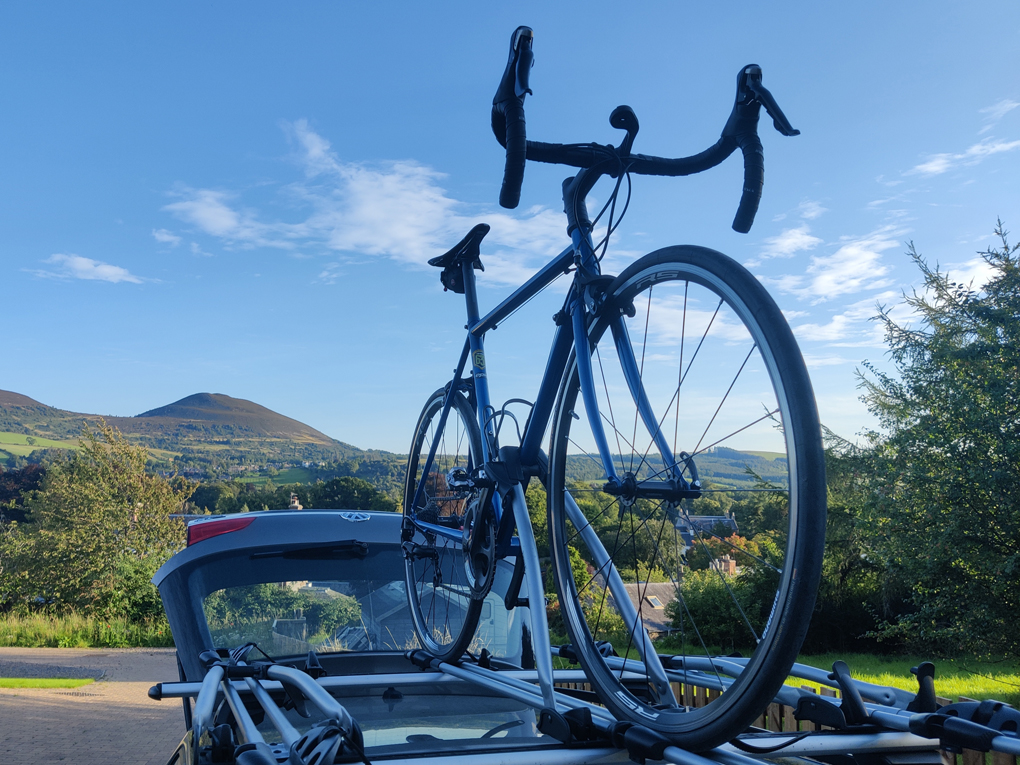 A blue racing bike is perched on the roof of a car, on a sunny morning with rolling hills in the background.