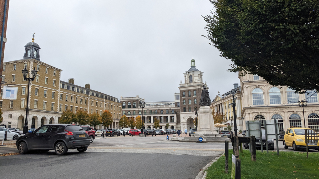 A town square. Edwardian era buildings surround car park and a statue of The Queen Mother.