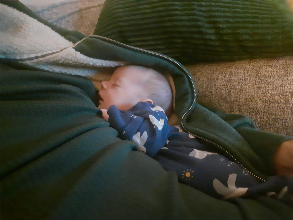 A newborn baby leaning tucked into the side of an open hooded jumper looking cosy.