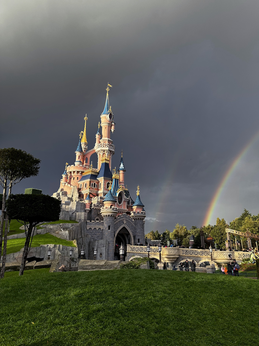 A double rainbow appears next to Sleeping Beauty's castle in Disneyland Paris, set against dark clouds with bright green grass in the foreground