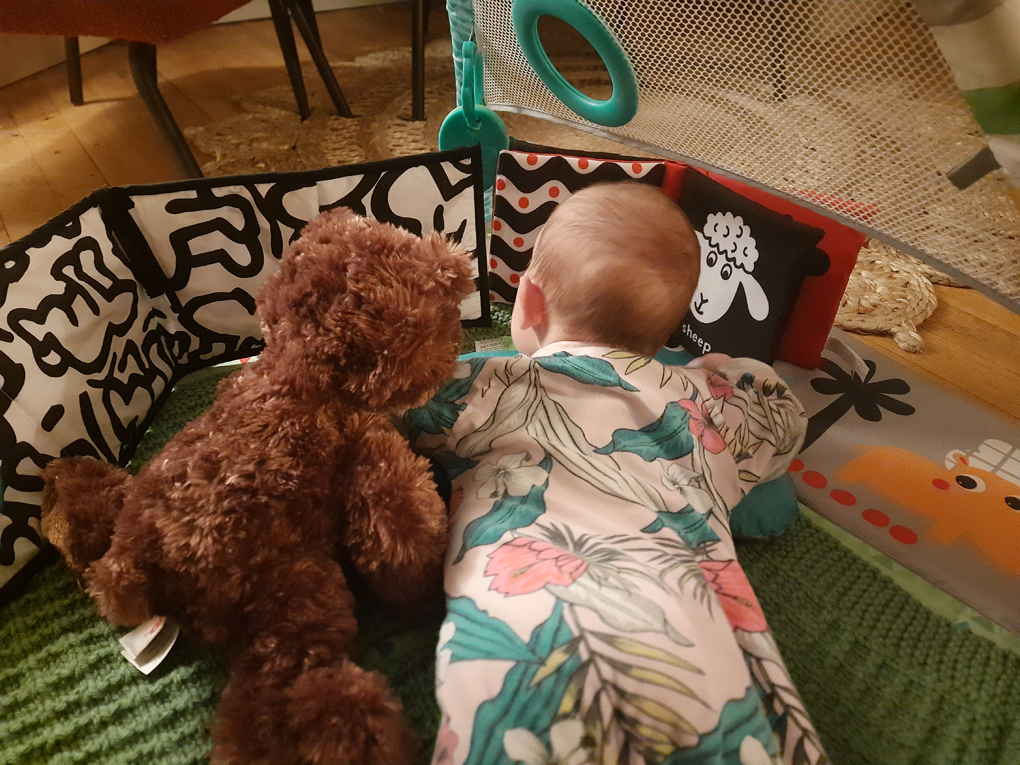 A baby lays on their tummy next to a stuffed bear about the same size as them. They gaze at black and white pictures together