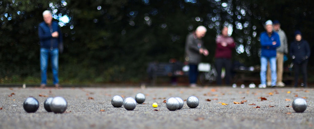 The image is take low down looking over the pitch towards the players. It is focused on the yellow jack with the silver pétanque balls around it. The players are in the far background, out of focus.