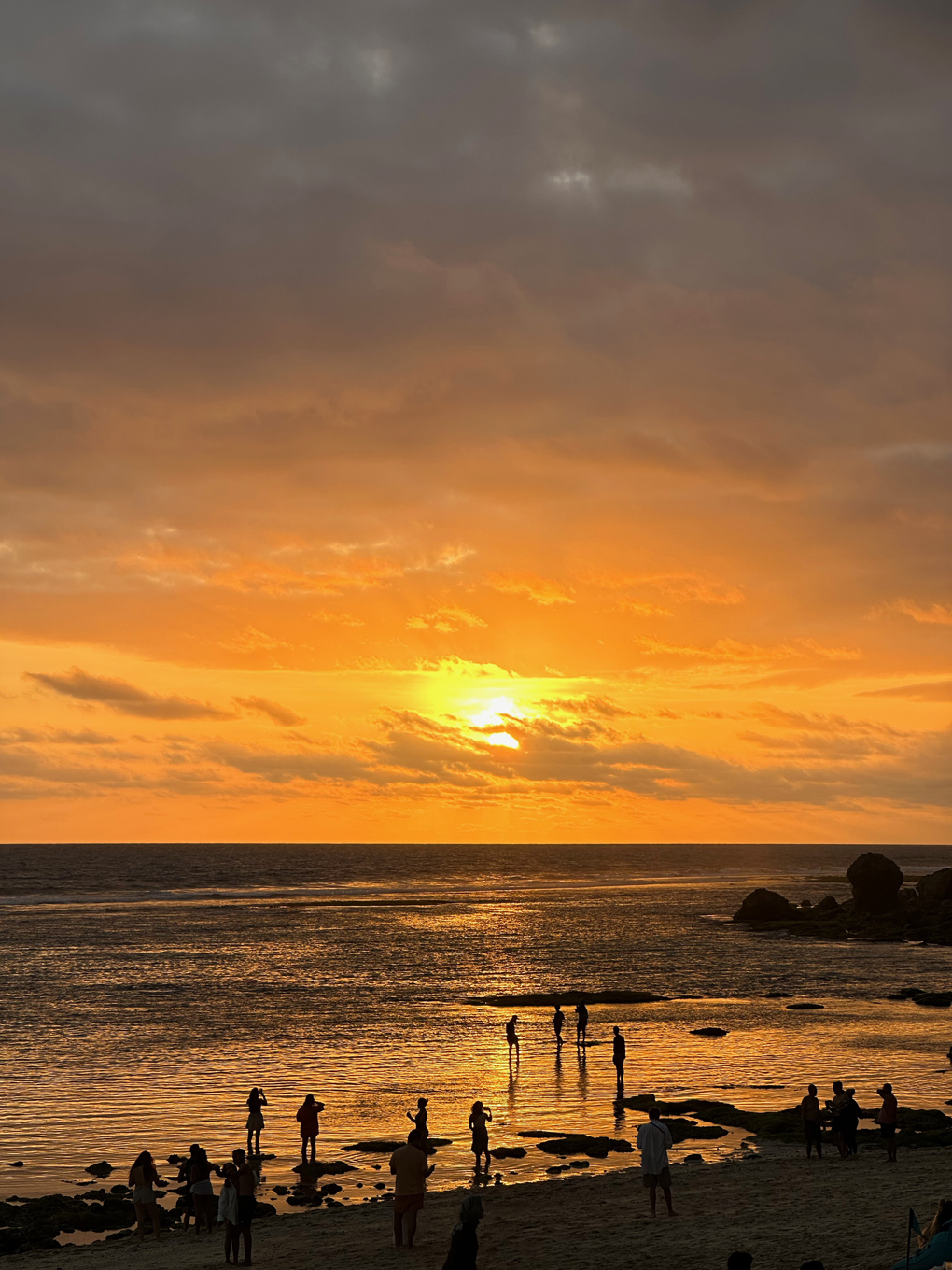 Orange sunset with some clouds banding. Open water and beach at bottom with people silhouetted as small figures.