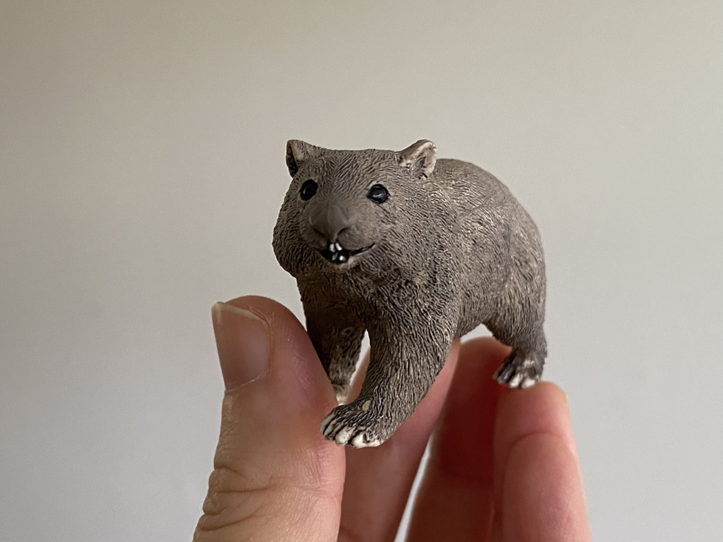 A small plastic wombat held aloft on fingertips. The wombat is smiling.