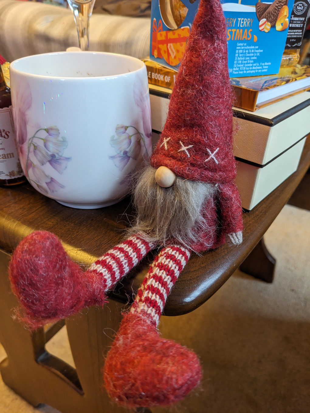 A felt bearded dwarf, dressed in red, sat on a table next to a mug