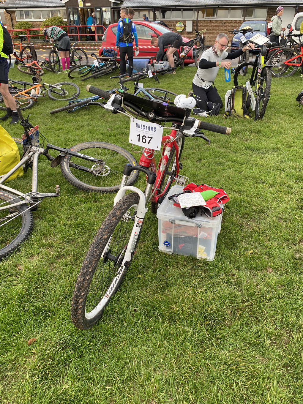 A mountain bike with a map board and attached race number (167) propped up against a box, in a field with other bikes.