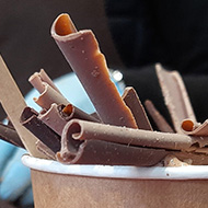 A cup of hot chocolate piled high with white milk, and dark chocolate shavings. In the background is a another large pile of chocolate.