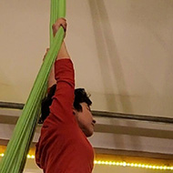 A short haired female holds onto a green silk hanging from the ceiling, one foot is wrapped up by the silk, the other leg is held forward in an attempt to be elegant. A mirror shows her reflection in the background