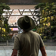 A man in an olive coloured t-shirt and blue shorts with a green felt hat photographed from behind. The inside of a domed structure ahead with an inactive water fountain.