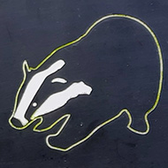 A metal sign attached to a wrought iron gate. The sign reads 'Keep the badgers off the lawns! Please close this gate between duck and dawn' with a picture of a badger in the centre. The gate is ajar leading to a lush green meadow behind it.