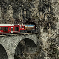 The front of a train going over a viaduct into a tunnel through a mountain in the alps. Taken from the back of the train.