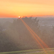 A thin orange, yellow, red sun beam strikes a park bench. A weak yellow beam is fading as the sun has nearly disappeared beyond the horizon. The horizon consists of a row of rooftops and trees in the distance
