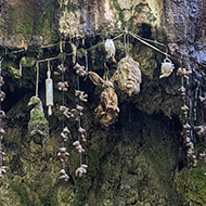 A rocky outcrop over which water drips into a pool below. A multitude of objects are hung in the path of the water as it drips down slowly turning them into stone including teddy bears, shoes, handbags and what looks like a human head