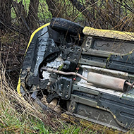 A rolled car off the road, on its right side with trees behind