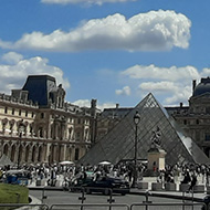 A wide view of the Louvre in Paris in the sunshine. The sky is vibrant blue with few clouds, the plaza is bustling with people, and the famous pyramid is in the centre.