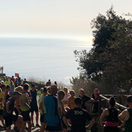 A crowd of people in running gear, with a stone fort on the left and the blue sky and sea extending to the horizon.