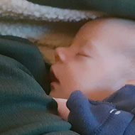 A newborn baby leaning tucked into the side of an open hooded jumper looking cosy.