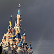 A double rainbow appears next to Sleeping Beauty's castle in Disneyland Paris, set against dark clouds with bright green grass in the foreground