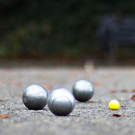 The image is take low down looking over the pitch towards the players. It is focused on the yellow jack with the silver pétanque balls around it. The players are in the far background, out of focus.