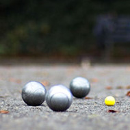 The image is take low down looking over the pitch towards the players. It is focused on the yellow jack with the silver pétanque balls around it. The players are in the far background, out of focus.