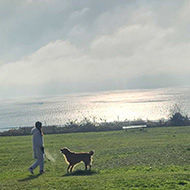 A lady and a dog stand on a patch of green. Behind is the sea. It is cloudy but sun rays peek through the cloud and the sun reflects a brilliant yellow off the bright blue sea