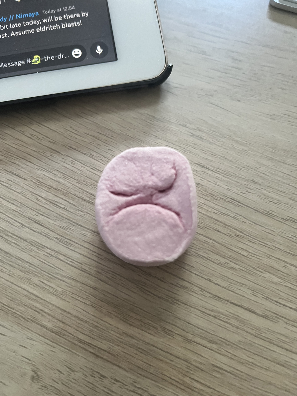 A pink marshmallow sat on a wooden office desk. The marshmallow has a grumpy face.