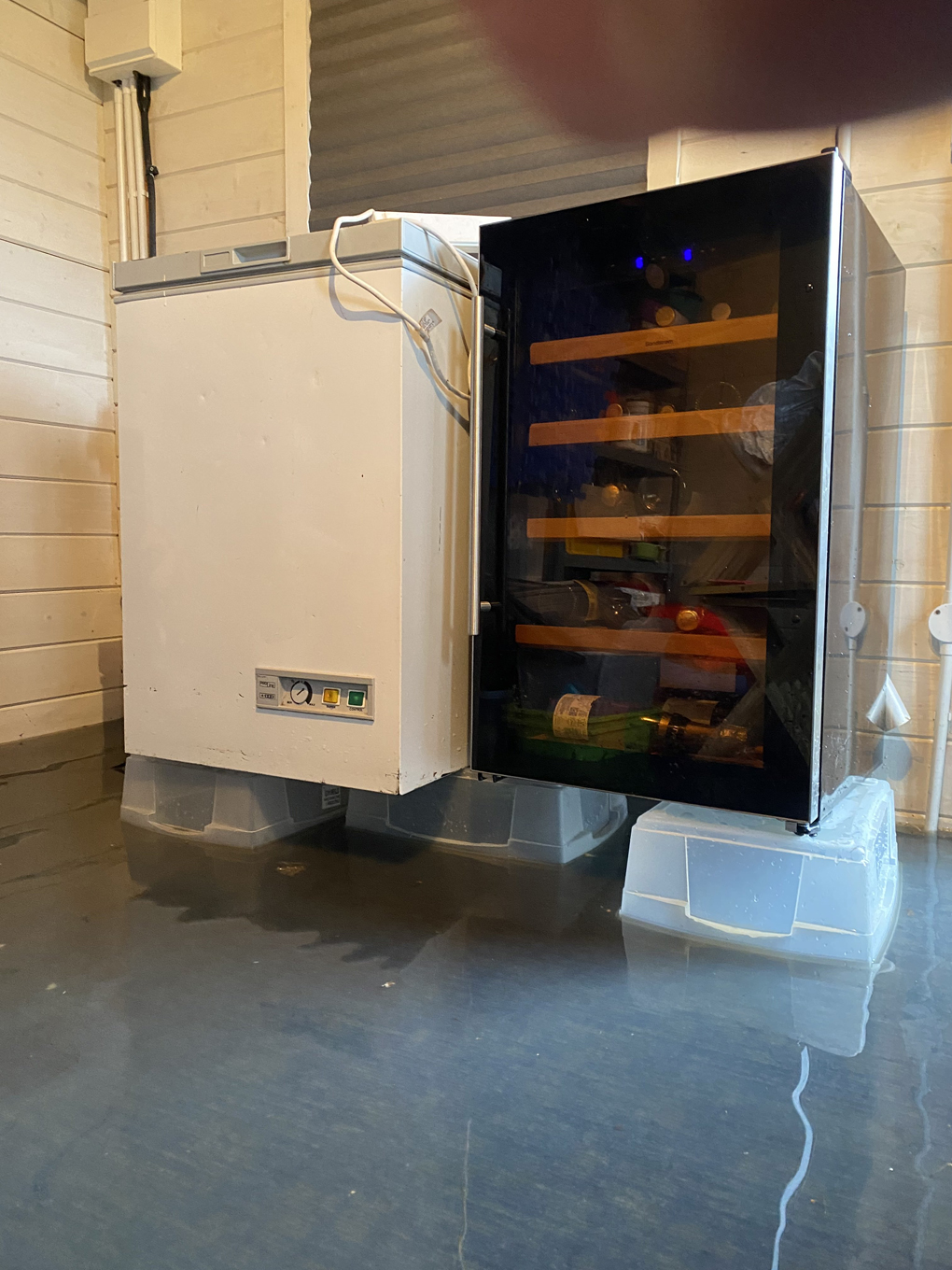 A small chest freezer and a wine fridge placed on top of plastic boxes to keep the white goods out of two inches of flood water in a log cabin.