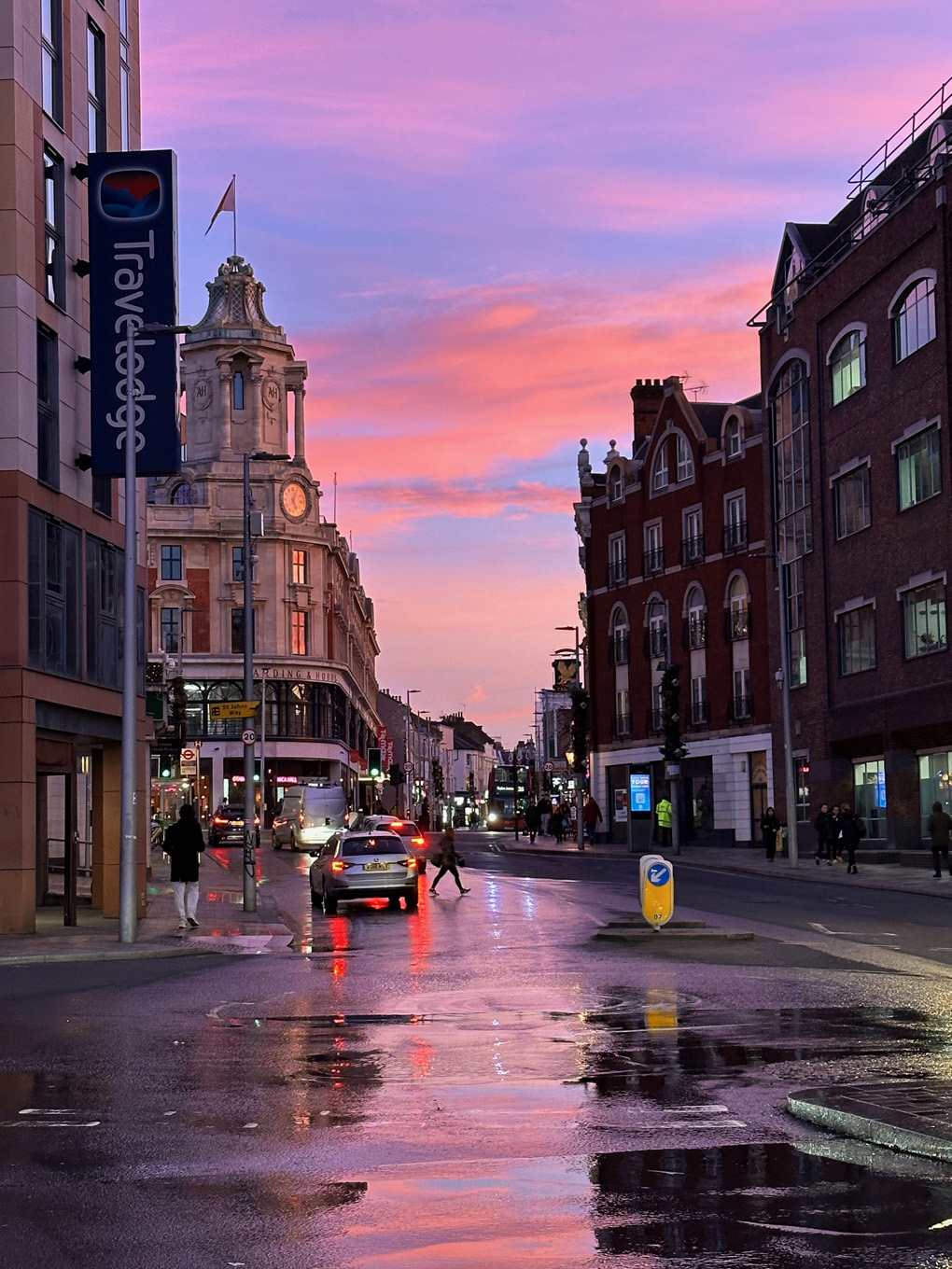 View down into Clapham Junction centre with some light traffic. Pink sunset reflected in large puddles on road.