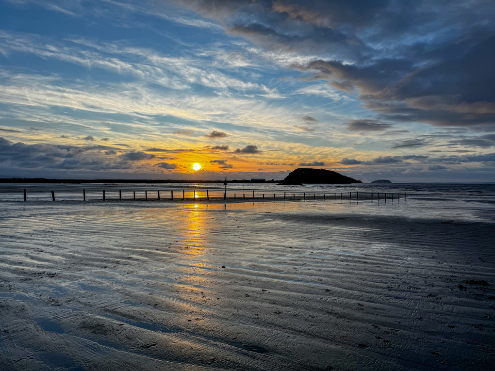 A bright golden orange sun beginning to set above a row of groynes protruding from wet sand flats