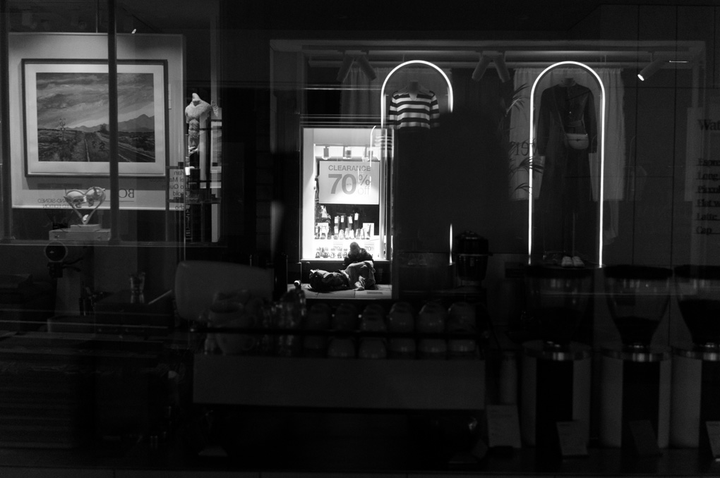 a homeless person sitting on the street in front of the bright window of a closed shop advertising a clearance sale