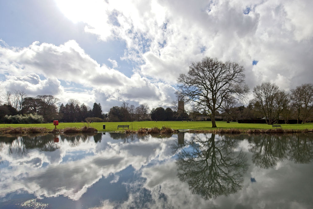 With a still lake in the foreground the sky, with streaks of white clouds, is reflected perfectly in the water. The composition is enhanced by the leafless tree on the far lake side, also reflected in the lake. In the distance the tower of the Cirencester Parish Church stands out.