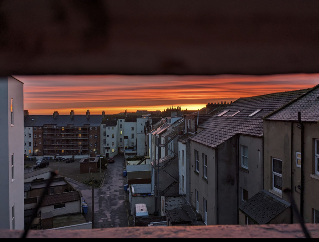 View of alleyway and rooftops. Whitby Abbey in the background shadowed against an orange and golden sunrise