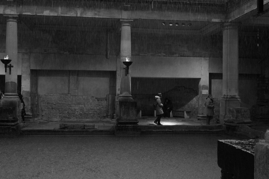 A lone woman steps into a pool of light in the Roman Baths, while in the foreground rain is falling into the main bath's water