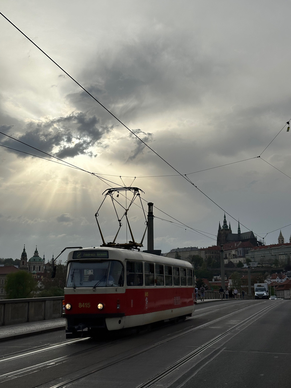 A Tatra T3 tram with Prague Castle in the background. Crossing over bridge with sun rays breaking out from behind clouds.