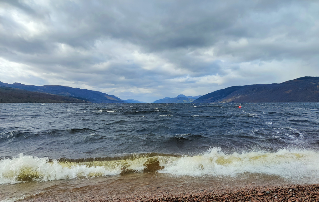 A wave crashes on the stones at the head of loch ness, looking down the loch, into the wind, hills rising beside the water and into the far distance under a brooding sky