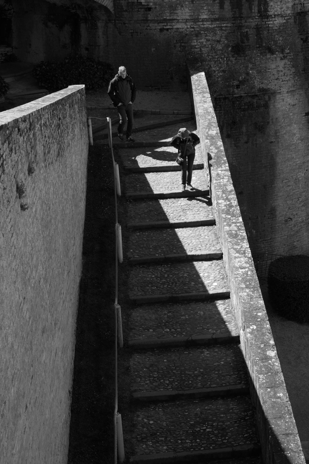 A black and white photo of two tourists walking down old stars, one framed in a sharply defined triangle of light