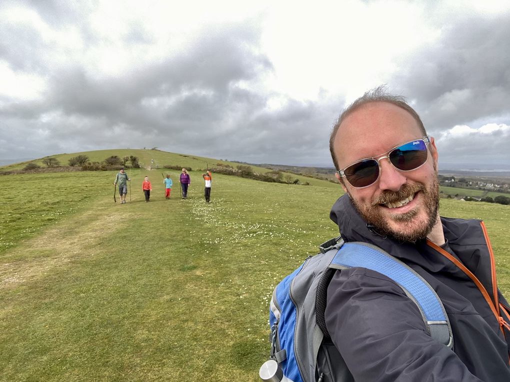 A group of children and adults in the background, walking on a green hill, with a bearded, smiling man in the foreground.